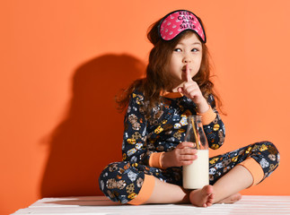 Little kid girl in pyjamas with kittens print is sitting on the floor with her legs crossed Turkish fashion with a bottle of milk showing shh sign