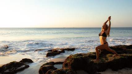 Fototapeta na wymiar Young woman practicing yoga, standing in Virabhadrasana pose on the beach. Sunset time. Tegal Wangi beach, Bali