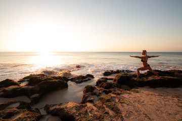 Young woman practicing yoga, standing in Virabhadrasana pose on the beach. Sunset time. Tegal Wangi...