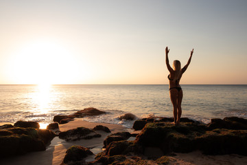 Fototapeta premium Excited young woman raising arms at the beach in front of the ocean. View from back. Sunset at the beach. Bali, Indonesia.