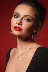 Closeup fashion vogue style portrait of young elegant woman with hairstyle and makeup. Girl with red lips wearing earrings and necklace. Studio shot against red background