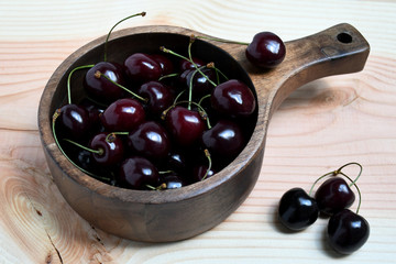 An image of a group of cherries in a wooden deep stewing pan, one berry on it and three cherries nearby on a light colored wooden surface