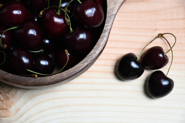An image of a fragment of a group of cherries in a wooden deep stewing pan, one berry on it and three cherries nearby on a light colored wooden surface 