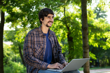 Young bearded brunet freelancer with mustache sitting with a laptop in a park. Work anywhere