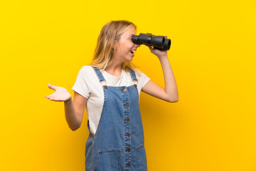 Blonde young woman over isolated yellow background with black binoculars