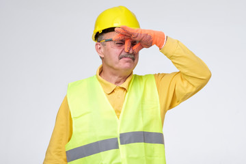 Displeased mature road worker in helmet and protective gloves closes nose because of unpleasant...
