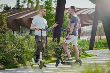 Two handsome men talking next to electric kick scooters at park landscape