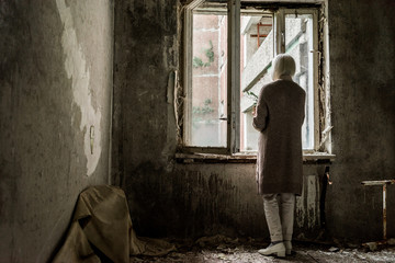 Fototapeta na wymiar back view of retired woman holding plant in empty room near windows