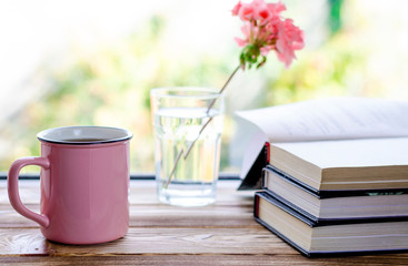 A pink cup with tea stands on a wooden windowsill next to books and a glass in which stands a pink geranium flower