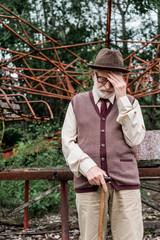 bearded retired man in hat and glasses standing with walking cane near damaged carousel