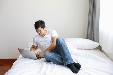 Fototapeta premium portrait of handsome caucasian in white shirt and blue jean using computer on bed in bedroom with relaxation and smiling face