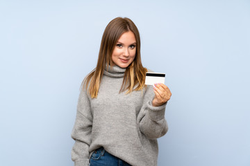 Fototapeta premium Teenager girl with sweater over isolated blue background holding a credit card