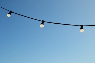 A string wire of outdoor light bulbs hanging against a clear blue sky. Light bulbs on the sky background. 