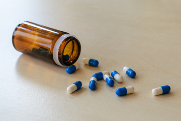 Scattered pills and brown glass jar on a table. White blue capsules spilled out of a glass pill bottle on a light wooden surface. To get sick, take pills or undergo treatment.