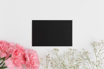 Top view of a black card mockup with gypsophila and a bouquet of pink carnations on a white table.