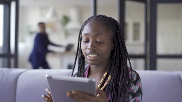 Young pretty African American woman with afro pigtails sitting at home working with her tablet. The girl calling her husband to show him picture. Leisure indoors