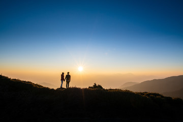 Obraz na płótnie Canvas Silhoutte of a couple hold hand of each other looking at sunset on top of hill with background of mountain and golden color of sunshine. Concept of teamwork, successful.