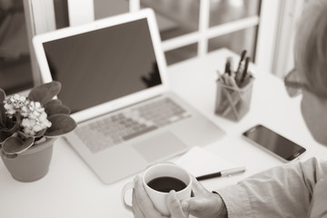 Hands of a senior woman holding a cup of coffee working outdoor in the terrace. Laptop and other devices close to her. Beautiful small flowering plant
