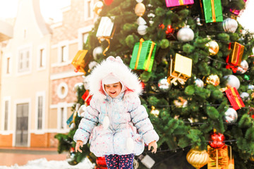 A little girl in a white hat and coat stands near a big Christmas tree. On the Christmas tree hangs a huge red ball. Festive winter photo.