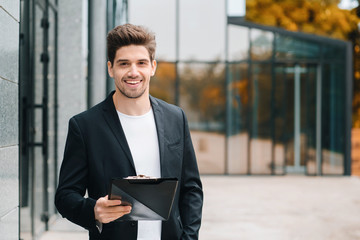 Portrait of young handsome man in jacket with documents, utility bills, report. Businessman near office building. He is satisfied with work of corporate staff