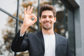 Successful happy man looking to camera and showing OK sign on office building background. Professional male manager wearing corporate suit jacket. Student guy outdoors