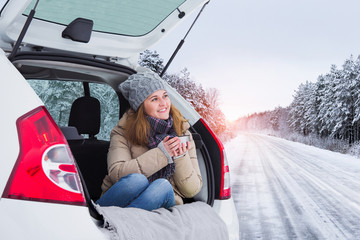 Woman in woolen hat sits in the trunk of the car and holds a cup of hot tea in her hands. 