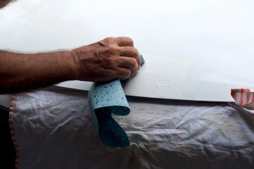 A man's hand is holding cloth rubbing area of white car hood that has paint damage. Hand is blurred from movement, paint damage in focus.