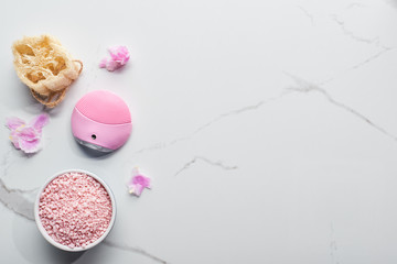 top view of silicone cleansing facial brush, bath salts and loofah on marble surface with pink petals