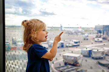 Fototapeta na wymiar Cute little toddler girl at the airport, traveling. Happy healthy child waiting near window and looking at planes. Family going on vacations by airplane. Canceled flight due to pilot strike.
