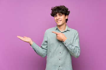 Young man over isolated purple wall holding copyspace imaginary on the palm to insert an ad