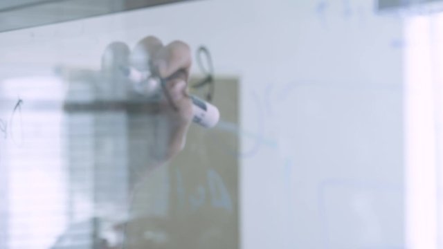 A teacher or a student writing math formulas on a transparent glass board with a black marker. Handheld panning shot.