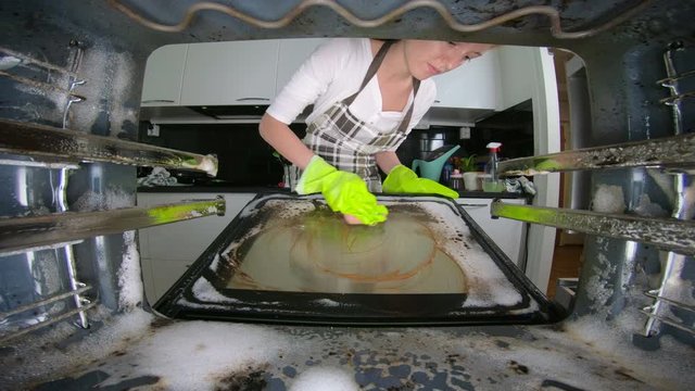 Woman housewife wearing green rubber gloves cleaning a dirty oven in the kitchen using a detergent.