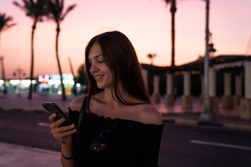 Fototapeta premium Happy brunette girl in black top reading message on her phone, blurred street with palms on the baclground