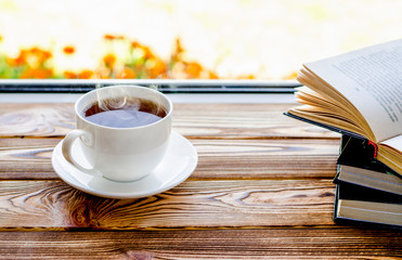 A white cup of tea stands on a wooden window sill next to books in the background of the window