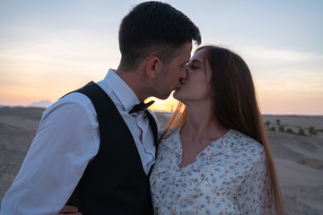 Beautiful young couple in elegant clothes kissing at sunset among the desert
