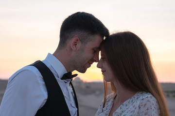 Romantic couple, brunette girl in delicate dress and handsome man in white shirt pressed their heads to each other at sunset among the desert