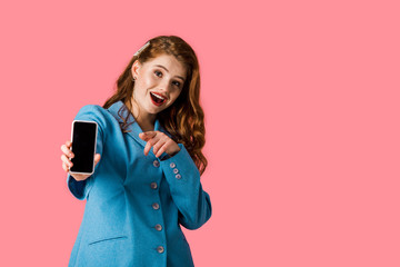 excited redhead girl holding smartphone with blank screen and pointing with finger isolated on pink