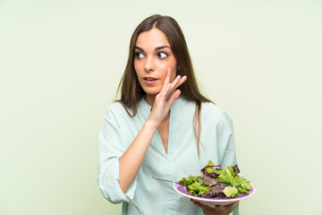 Young woman with salad over isolated green wall whispering something