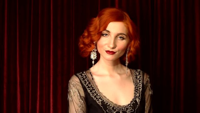 Retro woman with pale skin and red hair in black vintage dress dance against red background in theater. Vintage redhead woman with red lips, blue eyes and retro hairstyle. Beautiful silver earrings