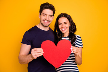 Photo of cheerful nice couple toothily smiling holding big red heart in love with each other while isolated with yellow background