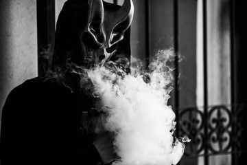 Vape man in devil mask and a hood smoking an electronic cigarette outdoors. Puffs of steam. Bad...