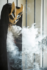 Vape man in devil mask and a hood smoking an electronic cigarette outdoors. Puffs of steam. Bad habit that is harmful to health. Vaping activity.