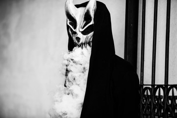 Vape man in devil mask and a hood smoking an electronic cigarette outdoors. Puffs of steam. Bad...