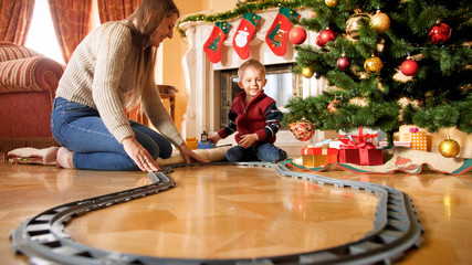 Cheerful smiling boy with young mother building railroad around Christmas tree at living room....