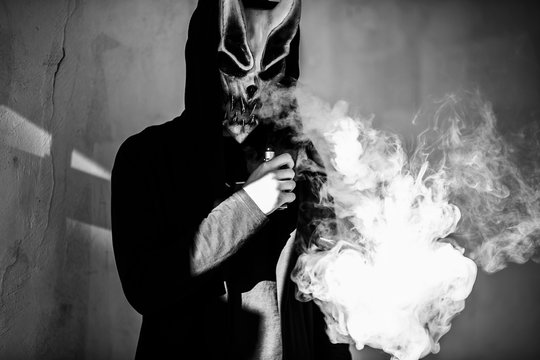 Vape man in devil mask and a hood smoking an electronic cigarette outdoors. Puffs of steam. Bad habit that is harmful to health. Vaping activity. Black and white.