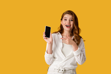 Naklejka premium happy redhead girl pointing with finger at smartphone with blank screen isolated on orange