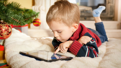 Naklejka premium Portrait of little boy lying on blanket next to Christmas tree and playing on digital tablet computer