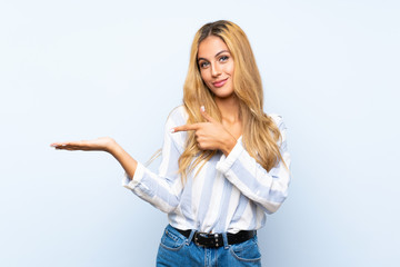 Young blonde woman over isolated blue background holding copyspace imaginary on the palm to insert...