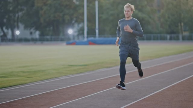 Athletic Fit Man in Grey Shirt and Shorts Jogging in the Stadium. He is Running Fast on a Warm Summer Afternoon. Athlete Doing His Routine Sports Practice. Slow Motion Tracking Shot.