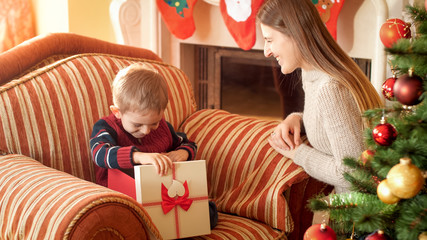 Photo of happy smiling little boy opens box with Christmas gift and looking inside. Mother giving gift to her child on New Year